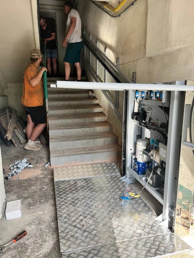 На зображенні представлено процес встановлення підйомника для маломобільних груп населення у під’їзді багатоповерхівки. Праворуч на сходах закріплено механізм підйомника з відкритою панеллю керування та електричними компонентами. Перед сходами розташована металева платформа з рифленою поверхнею. Троє робітників перебувають у процесі виконання монтажних робіт біля сходів, на яких видно поруччя та сходинки, оздоблені плиткою