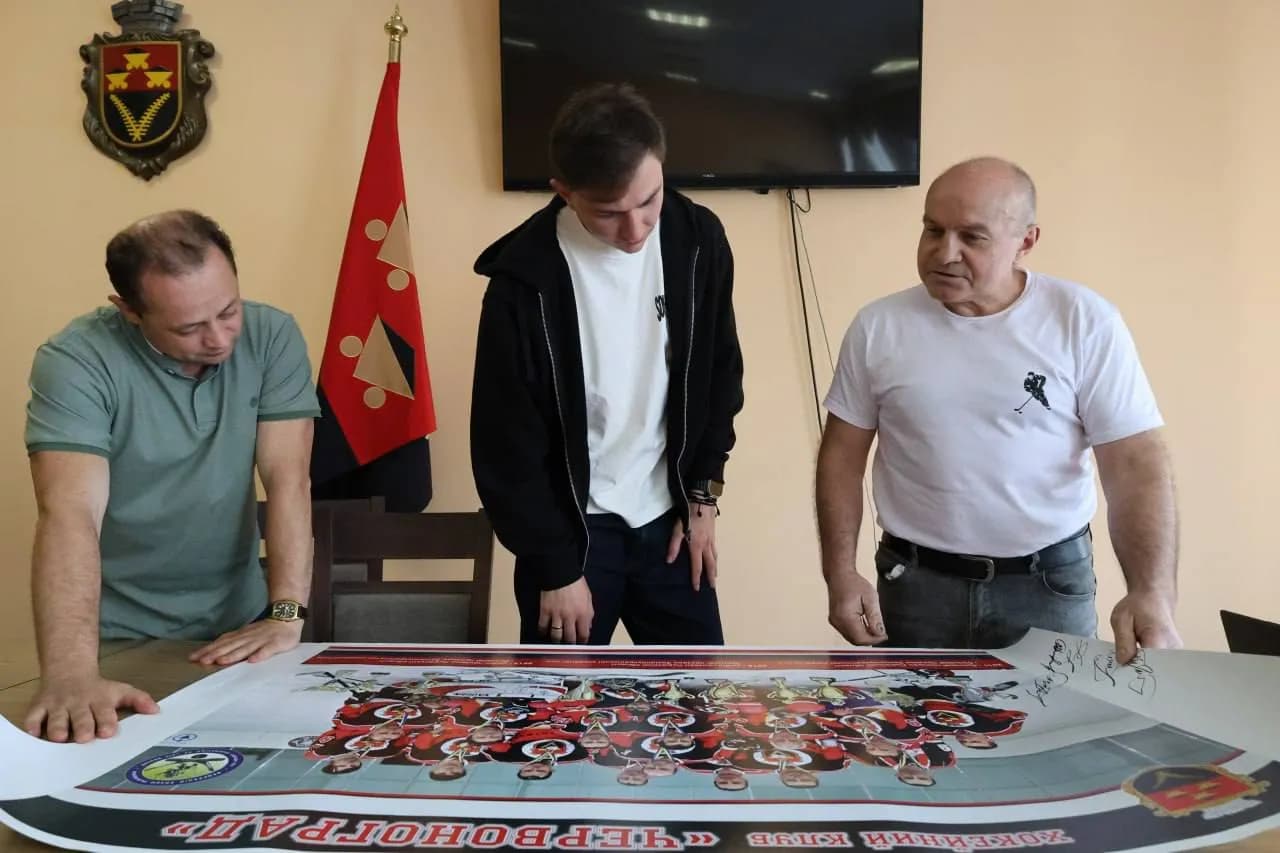Хокеїст, секретар ради, та президент хокейного клубу розглядають плакат, де зображена команда хокейного клкбк "Червоноград"