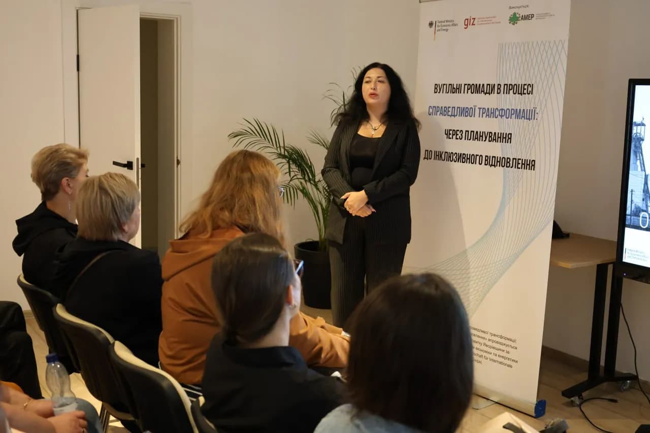 Жінка-лектор стоїть на фоні банера із надписом "Вугільні громади в процесі справедливої трансформації - через планування до інклюзивного відновлення"