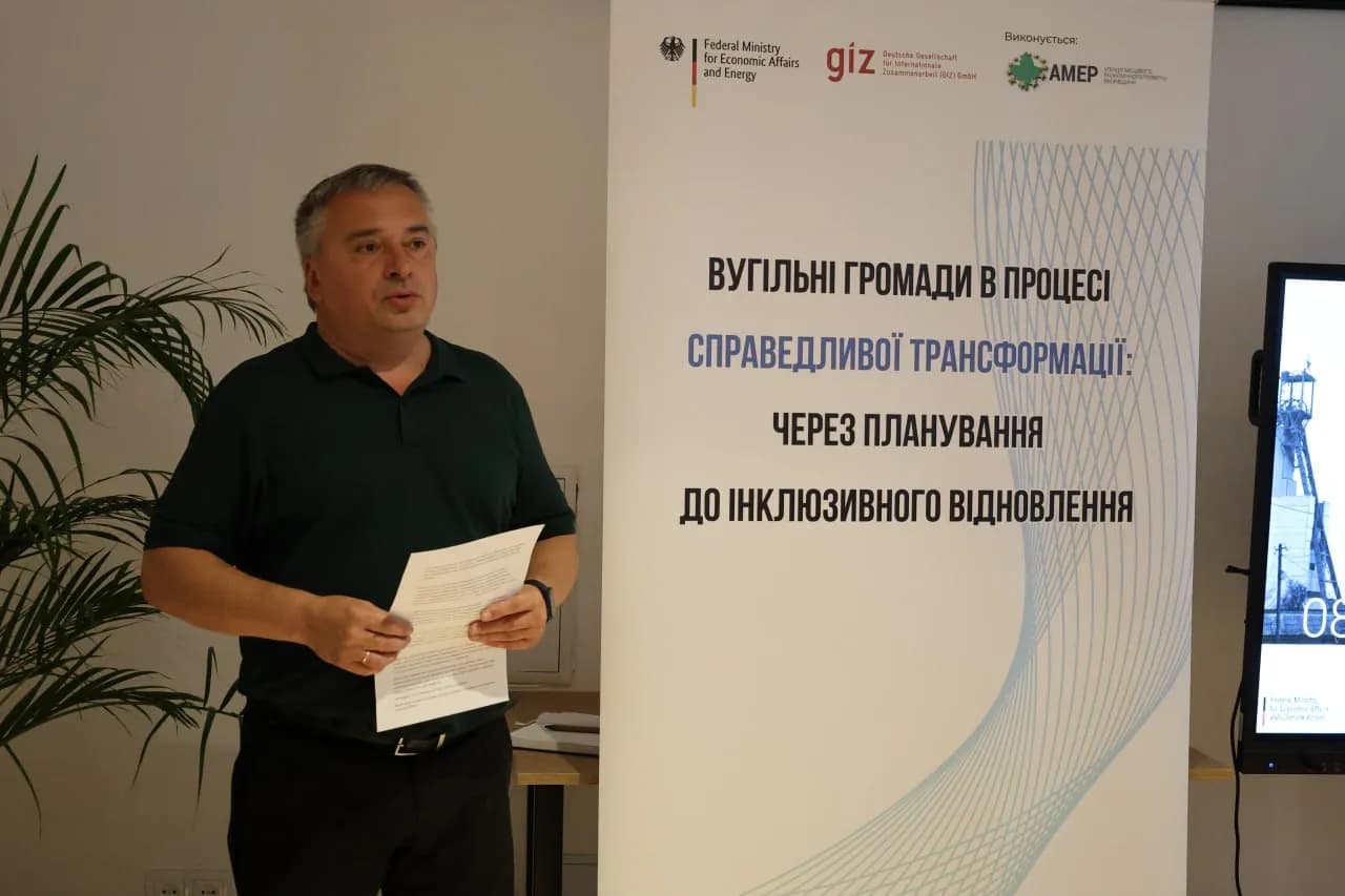 Чоловік-лектор стоїть на фоні банера із надписом "Вугільні громади в процесі справедливої трансформації - через планування до інклюзивного відновлення"
