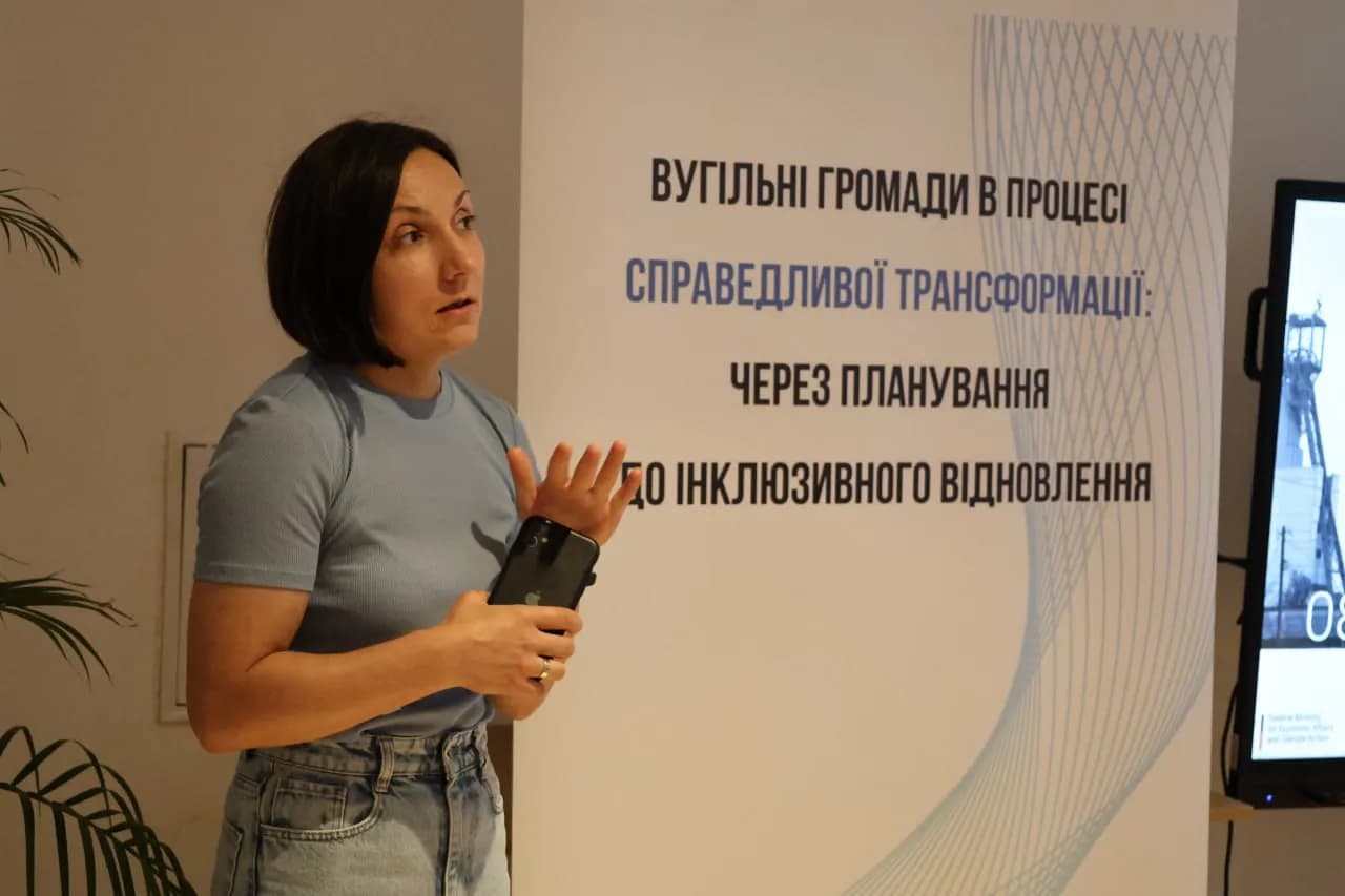 Жінка-лектор стоїть на фоні банера із надписом "Вугільні громади в процесі справедливої трансформації - через планування до інклюзивного відновлення"