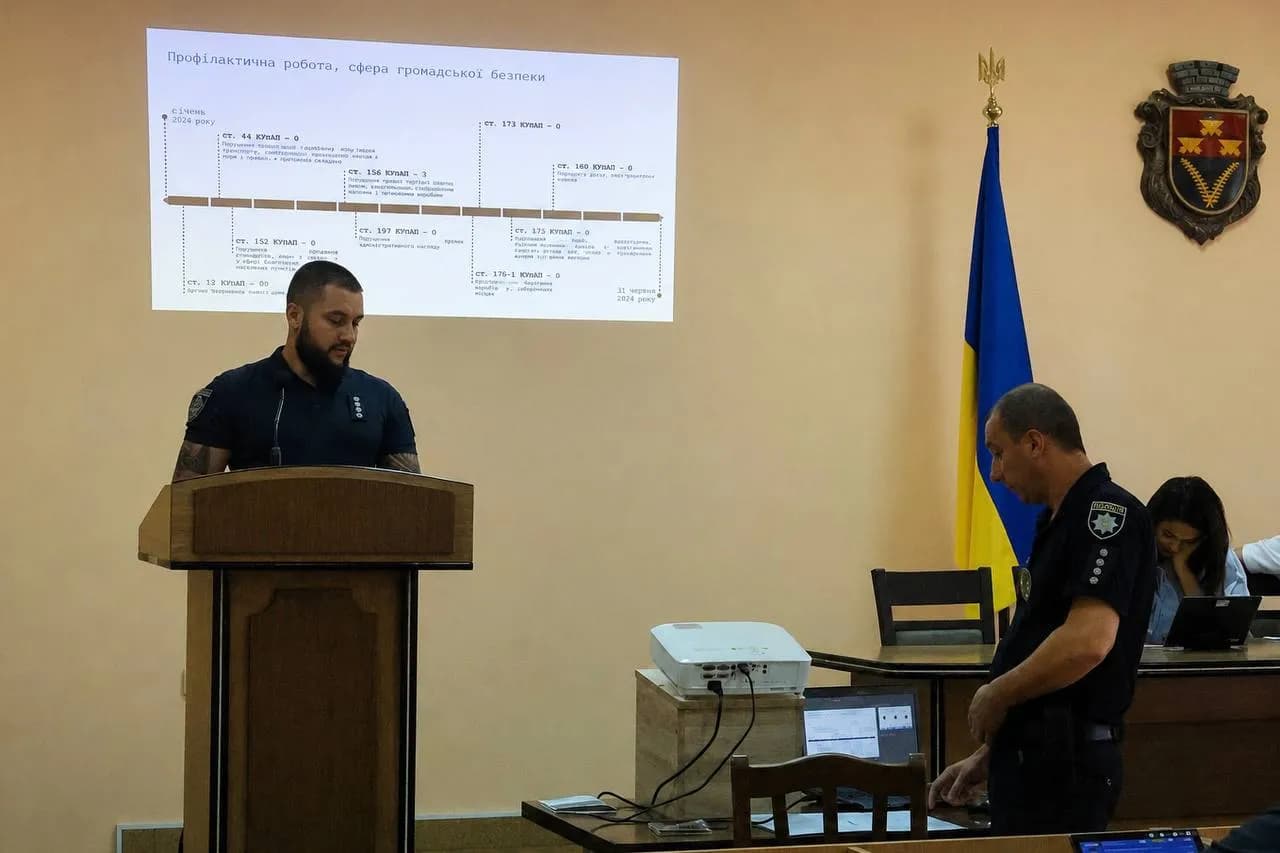 Поліцейський за трибуною звітує про роботу на фоні презентації, інший за ноутбуком перемикає слайди презентації
